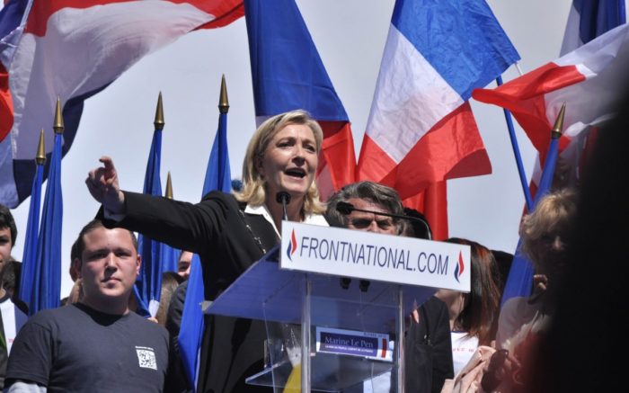 Marine Le Pen hinter einem Rednerpult. Hinter ihr sind französische Flaggen.