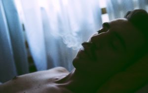 Ein junger Mann mit nacktem Oberkörper, der raucht, liegend im Halbschatten