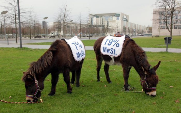 Zwei Esel mit je einer brschrifteten Satteldecke, auf der die Mehrwertsteuersätze 7 und 19 Prozent aufgedruckt sind, grasen auf einer Rasenfläche im Berliner Regierungsviertel. Im Hintergrund ist das Bundeskanzleramt zu sehen.