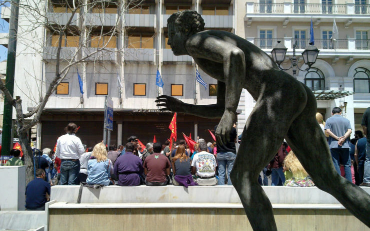 Skulptur eines Läufers im Vordergrund, im Hintergrund sitzende Menschen einer Straßenversammlung
