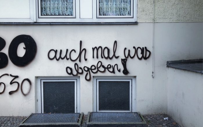 Schwarzes Graffiti auf der Wand: "auch mal was abgeben!" in Schreibschrift. darunter Kellerfenster, darüber Fenster mit Spotzen-Gardinen.