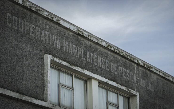 Auf einem grauen Gebäude steht der italienische Name einer Kooperative. Im Hintergrund sieht man den bewölkten Himmel.