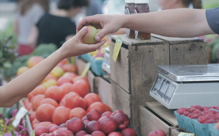 An einem Marktstand legt ein Verkäufer einen Apfel in die Hand eines Käufers.