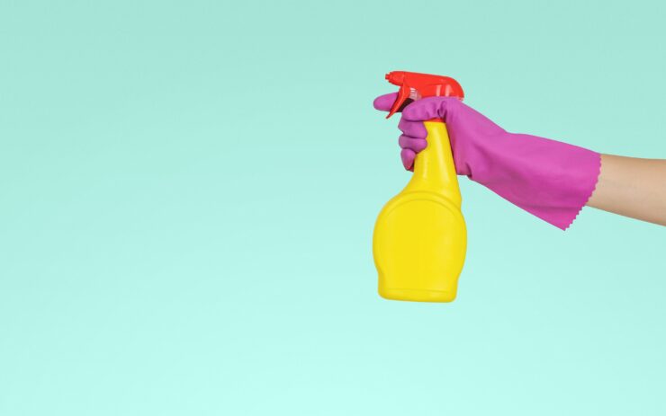Vor einem türkisem Hintergrund hält eine Hand mit einem Putzhandschuh eine Spritzflasche wie eine Pistole