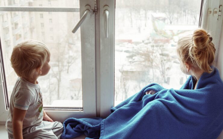 Zwei Kinder schauen aus dem Fenster auf eine schneebedeckte Stadt. Eins ist ein eine blaue Decke gewickelt.