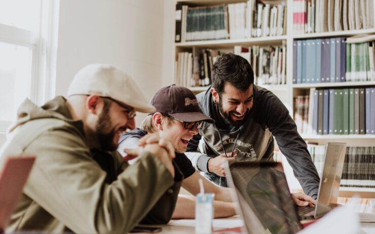 Drei Männer sitzen in typischer Atmosphäre von Start-ups vor Laptops und lachen