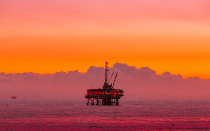 Ein roter Sonnenuntergang über dem Meer. Eine Förderplattform, wie Erdölkonzerne sie einsetzen, steht vor einer Wolke.