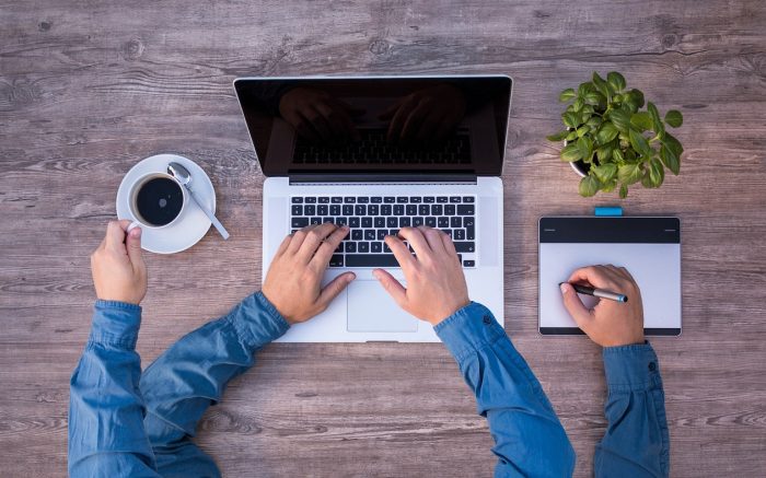 Vier Hände auf einem Schreibtisch arbeiten simultan an einem Laptop, halten eine Tasse Kaffee und schrieben Notizen.
