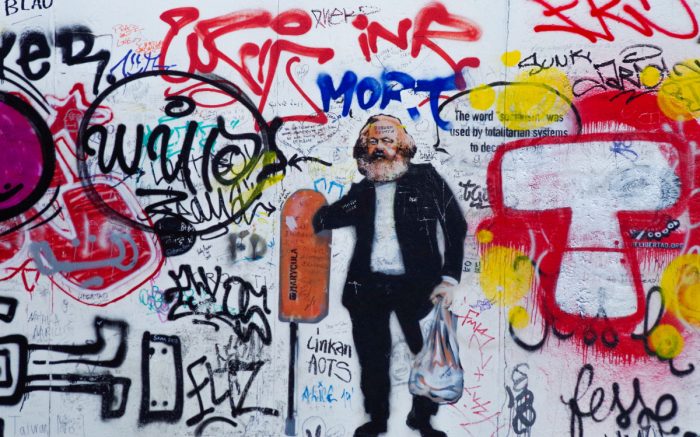 Eine Abbildung von Karl Marx als Grafitti zwischen anderen Graffitis. Er trägt einen Business causal Anzug und zieht Mehrwert aus einem Berliner Mülleimer.